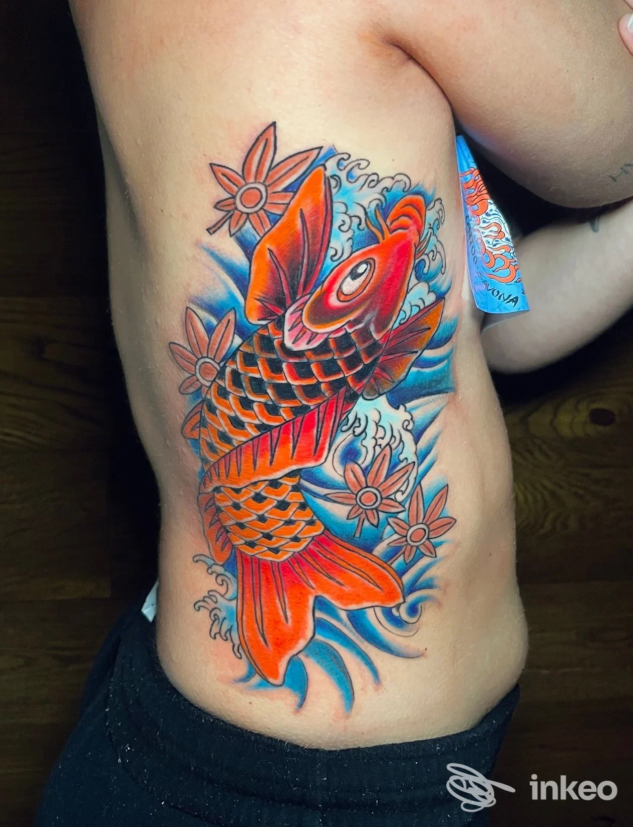 Tatuaggio carpa koi colorata con onde e fiori sul fianco, realizzato da Alien8 Tattoo Studio a Savona.