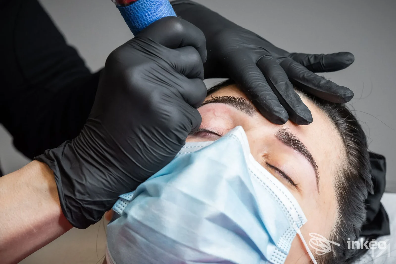 Trucco permanente alle sopracciglia in corso: cliente con occhi chiusi e mascherina, operatore con guanti neri nello studio MARINA AMBRIFI TRUCCO PERMANENTE, Latina.