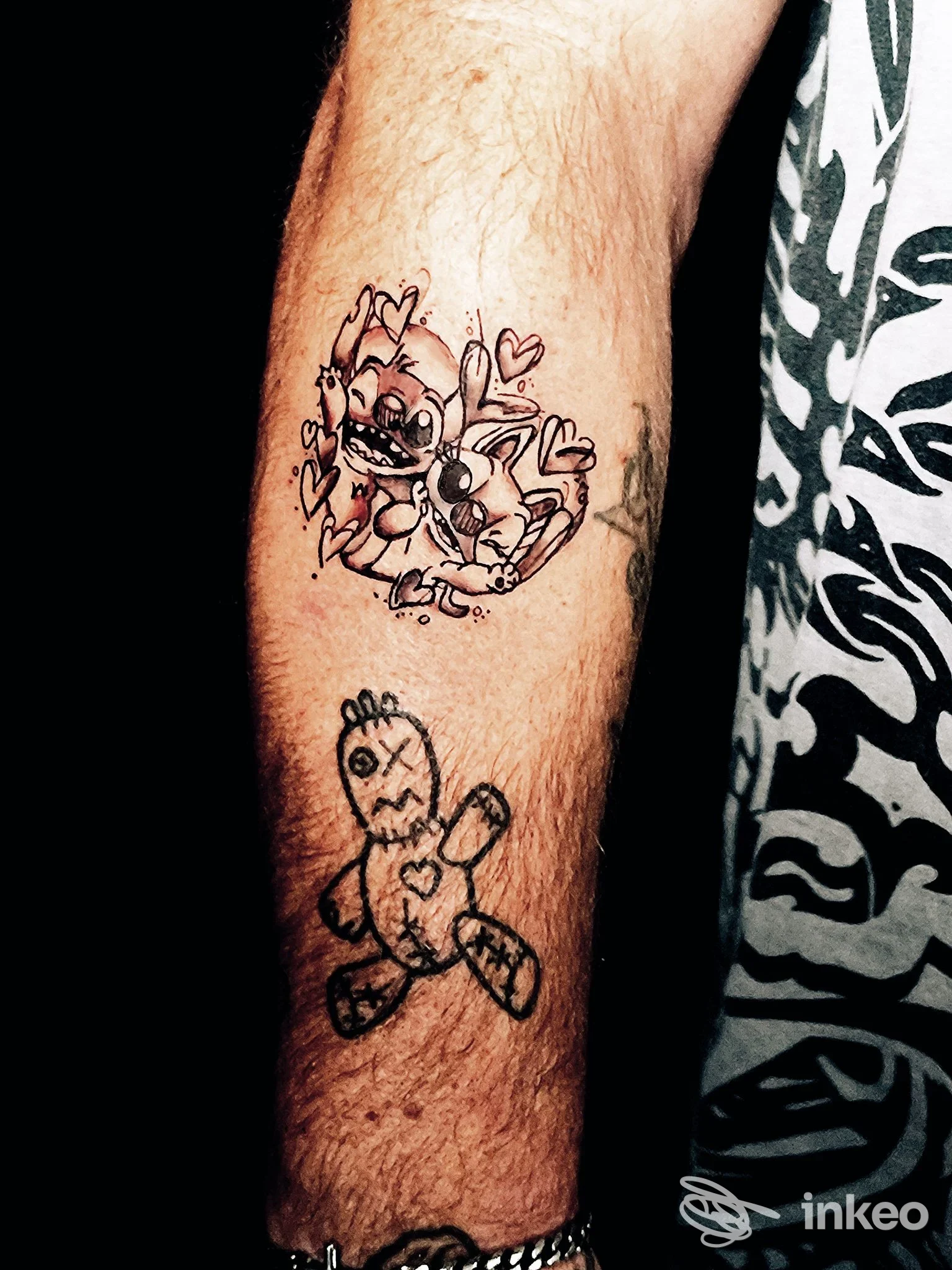 Avambraccio tatuato con due disegni in nero: personaggio cartoonesco con cuori e una bambola voodoo. Cerbero tattoo studio, Villafranca Piemonte.