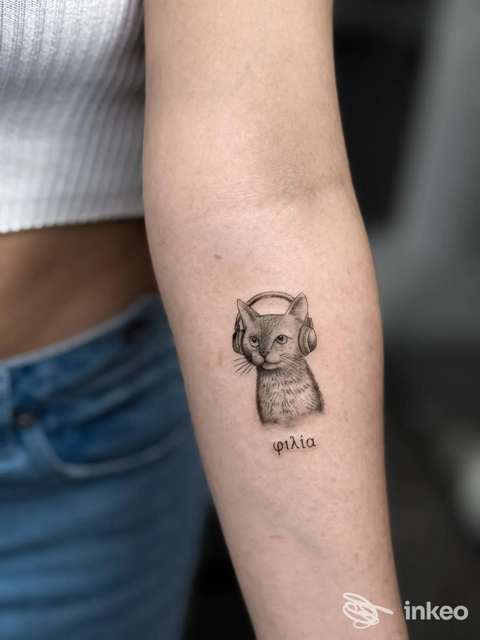 Tatuaggio di un gatto con cuffie sull'avambraccio, con la parola 'φιλία' sotto, realizzato da Pigmento Dermo Studio a Torino.