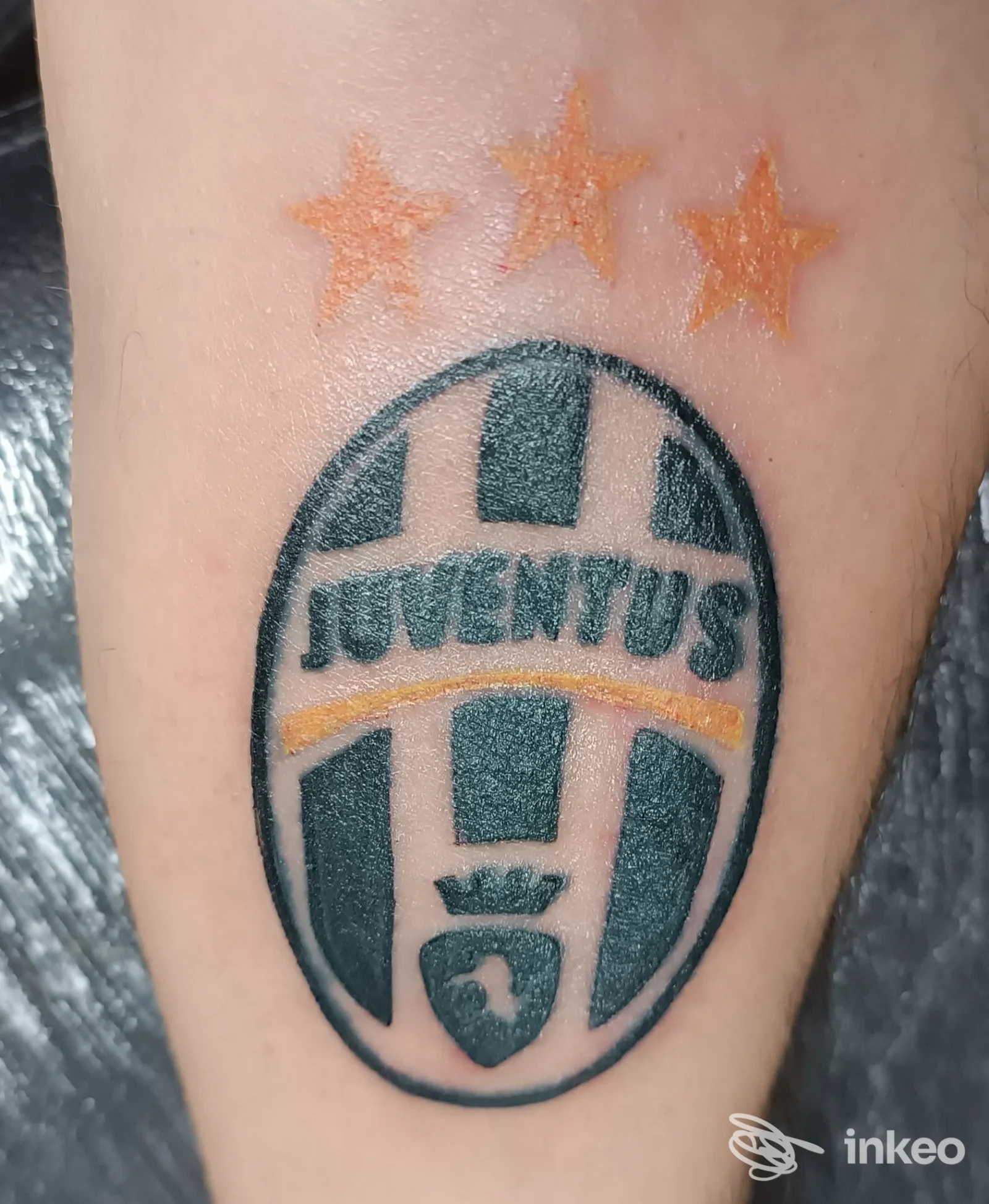 Tattoo realistico su pelle raffigurante un logo originale della Juventus in ovale, con tre stelle arancioni sopra, visibile su una porzione di avambraccio/pelle, colori neri e gialli.