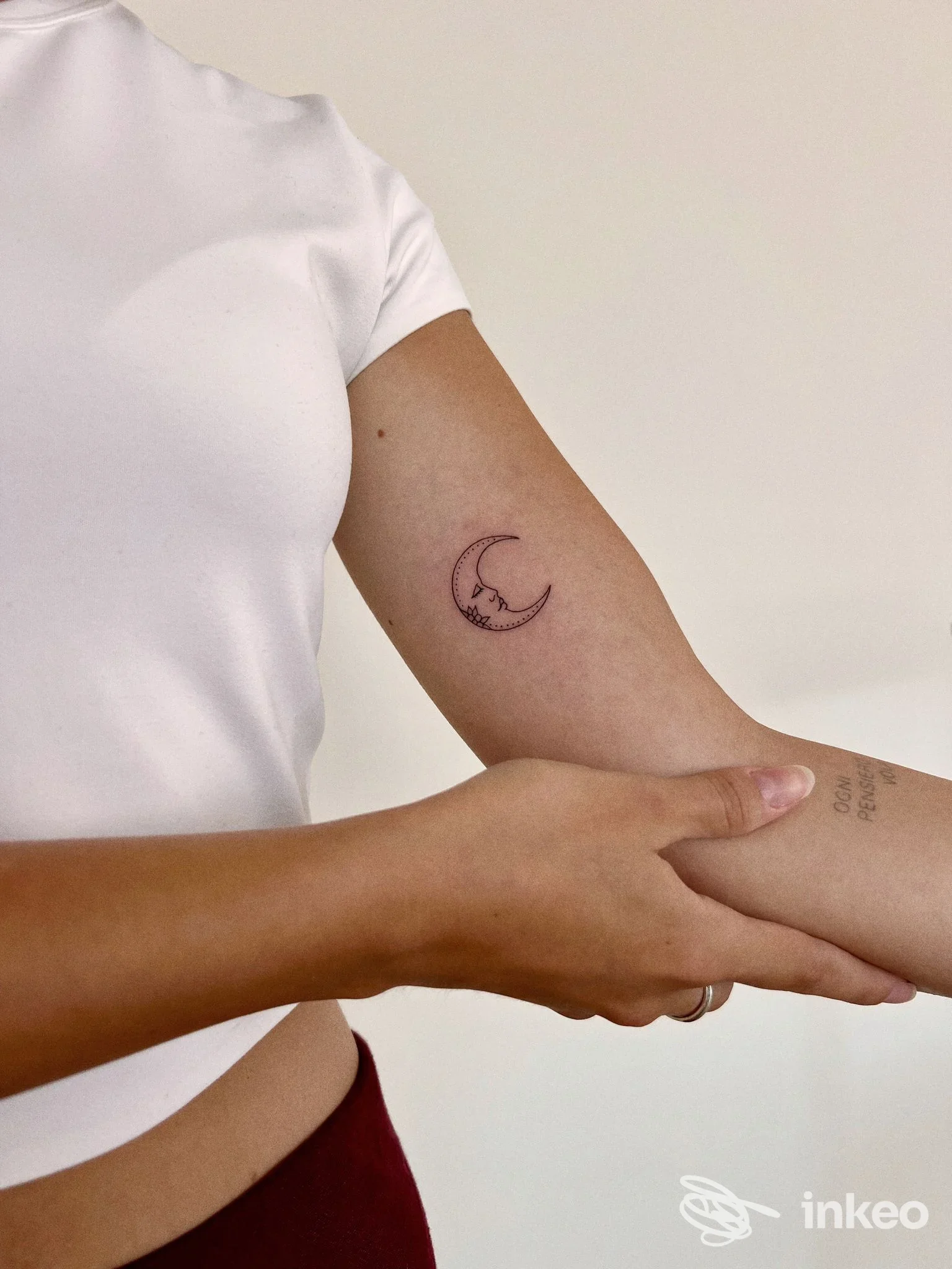 Tatuaggio a forma di mezzaluna su avambraccio di una persona, contorno sottile con sfondo neutro, maglietta bianca. Studio Fineline Tattoo Studio.