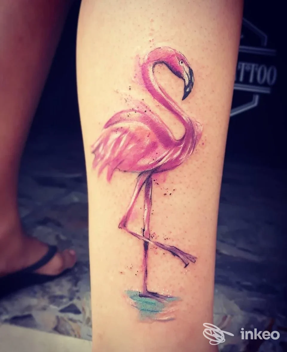 Tattoo realistico di un flamingo rosa sulla gamba di una persona, realizzato direttamente sulla pelle, con tonalità rosa e blu che riflettono sull’acqua.