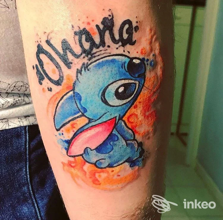Tatuaggio colorato stile acquerello su avambraccio realizzato da BarbieSlayer Tattoo, Macerata, rappresenta un personaggio cartoonesco blu e la scritta 'ohana'.