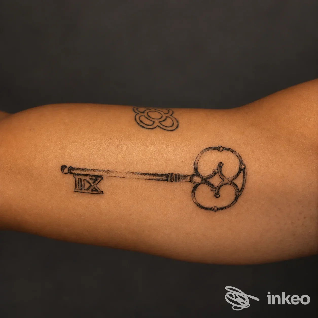 Tatuaggi neri su avambraccio: una chiave ornamentale e un piccolo fiore. Foto inviata da Be Unique Tattoo, Marotta.