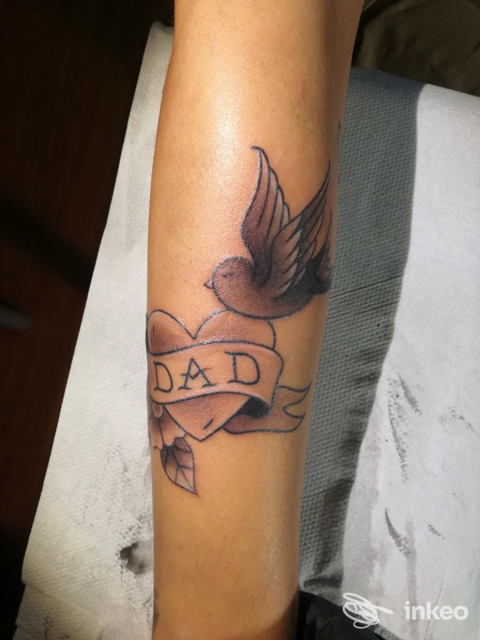 Avambraccio con tatuaggio a cuore e nastro "DAD" sormontato da una colomba, realizzato da DIABOLINK tattoo&piercing a Petritoli.