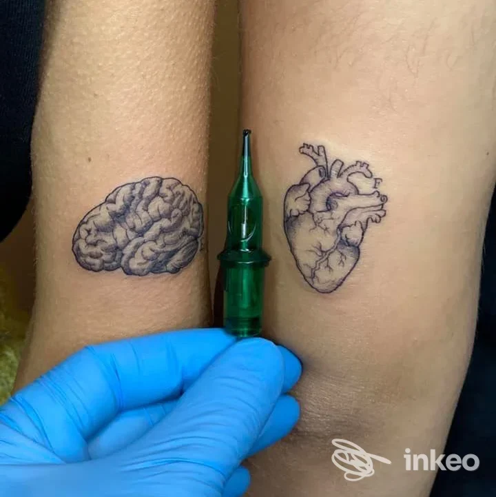 Due tatuaggi realistici su entrambe le avambraccia: cervello a sinistra e cuore anatomico a destra, con una pinza/tatuaggio in primo piano; nessun volto visibile.