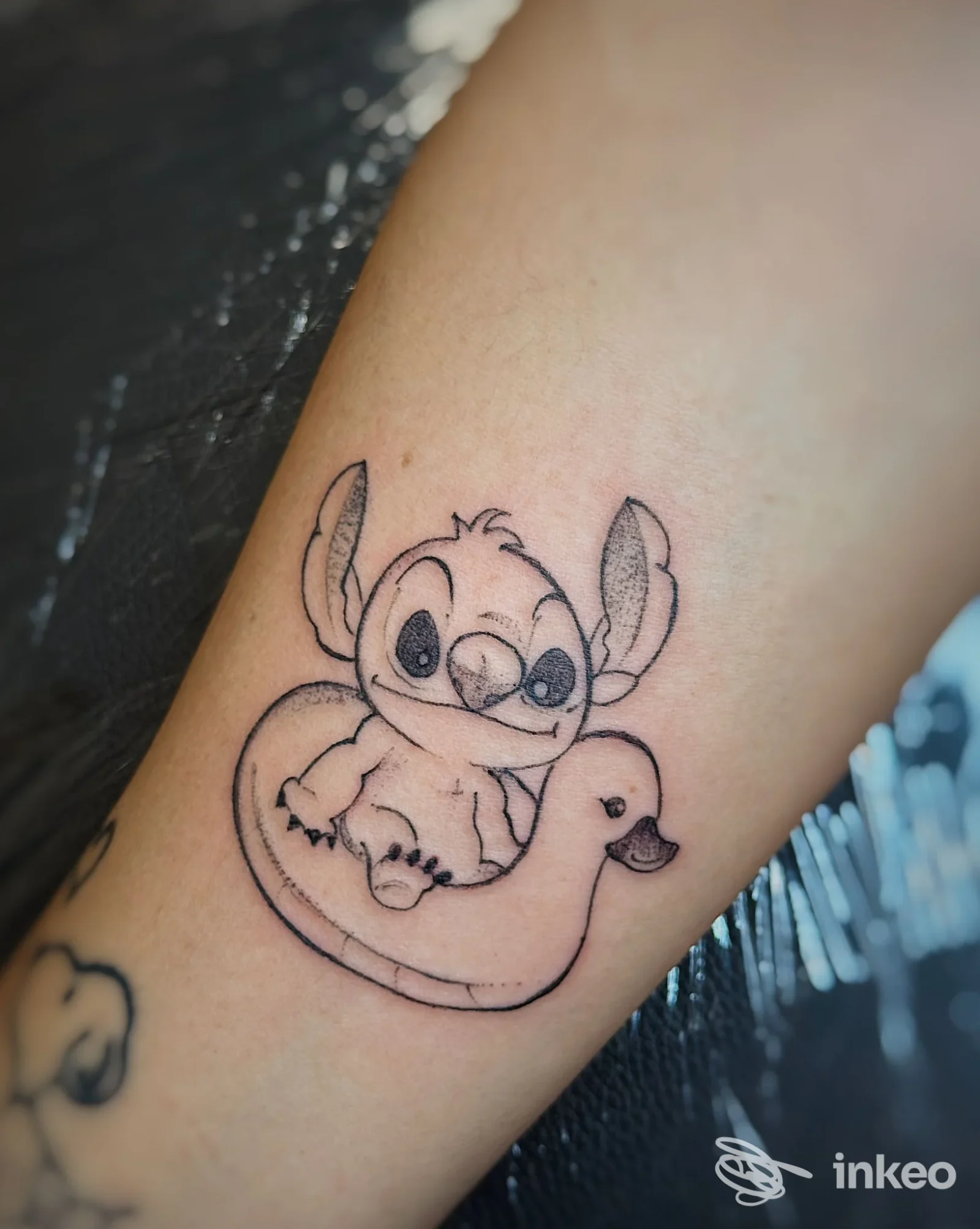 Tatuaggio linework di Stitch seduto su un gonfiabile a forma di anatra sull'avambraccio, realizzato da Gloria Tattoo a Camerino.