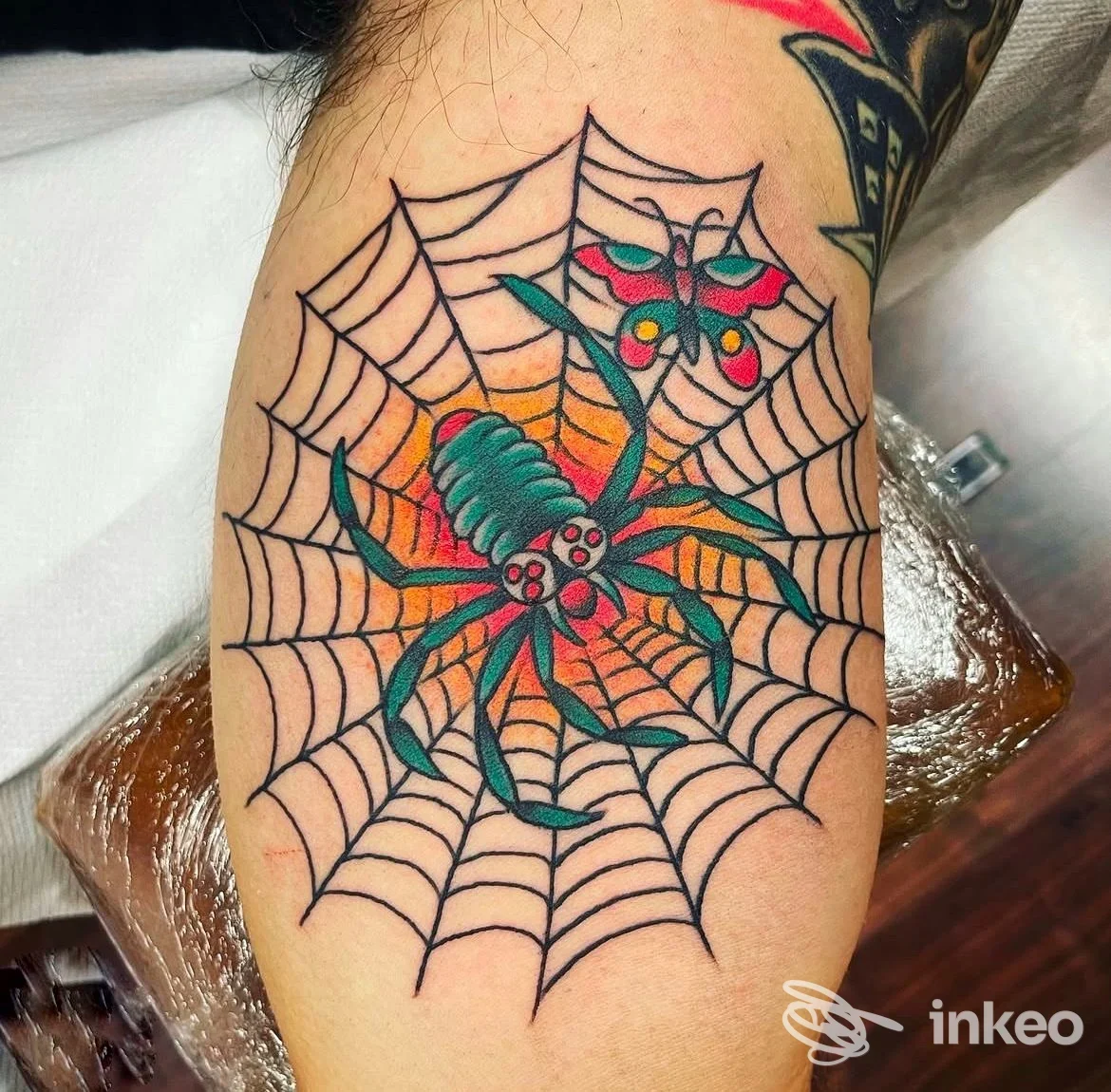 Tatuaggio tradizionale di un ragno verde su ragnatela con farfalla, scatto sull'avambraccio. Homeward Tattoo, Jesi.