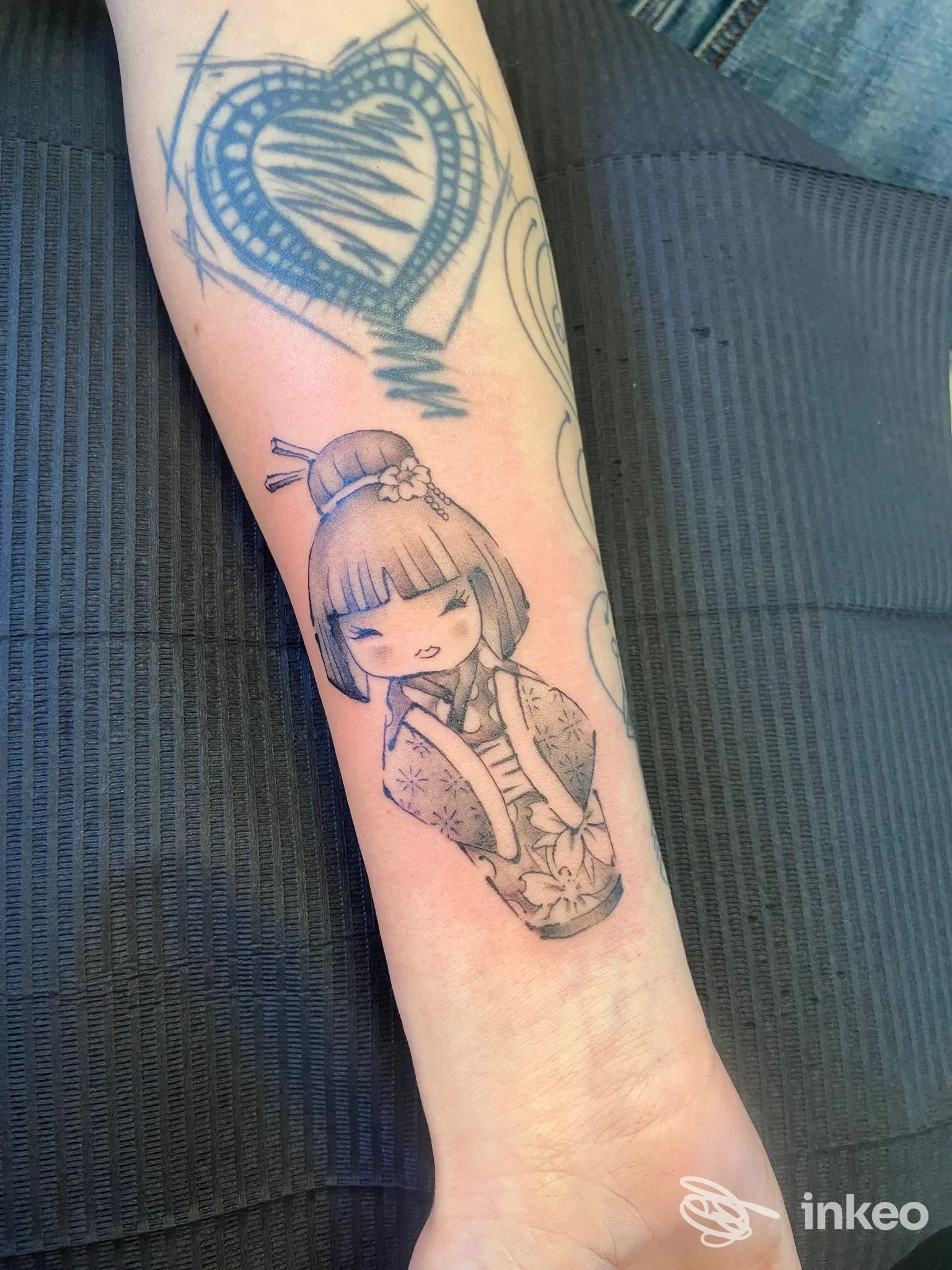 Primo piano su avambraccio con tatuaggio in nero e grigio di una bambola kokeshi e un cuore stilizzato, immagine fornita da Mary Skin - Tattoo & Piercing, Acqualagna.