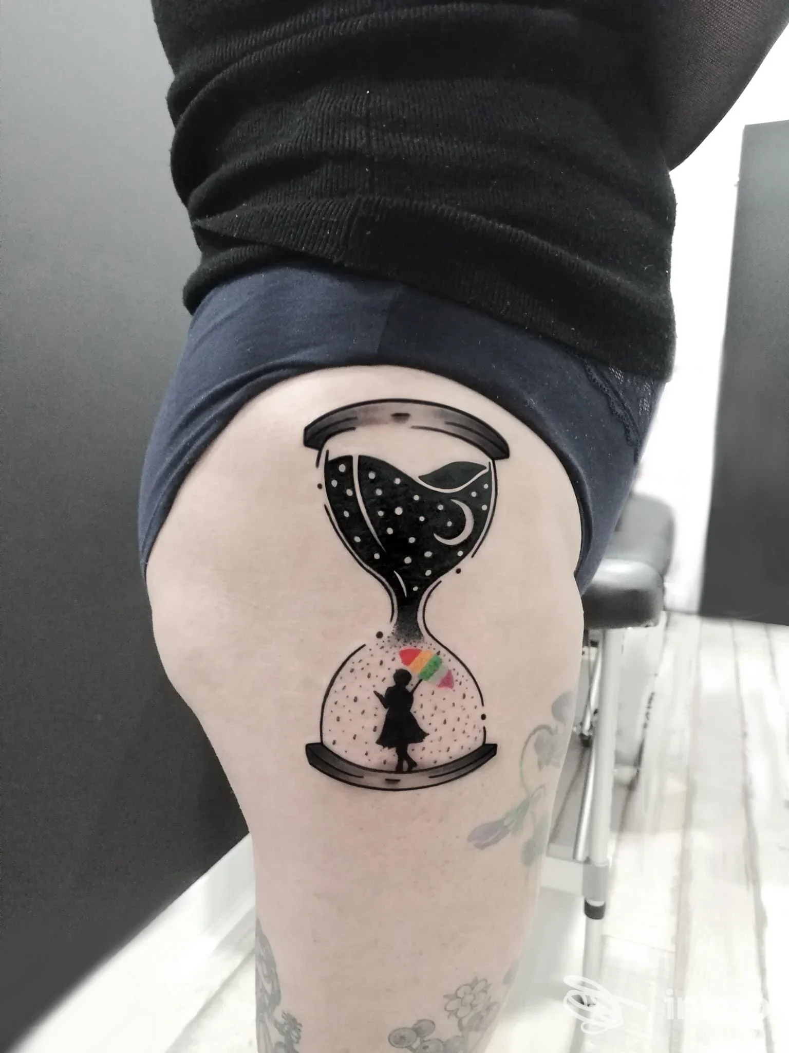 Tatuaggio a clessidra stilizzata sulla coscia con silhouette e piccoli dettagli colorati, realizzato da Nova Studio Tattoo & Piercing a Jesi.