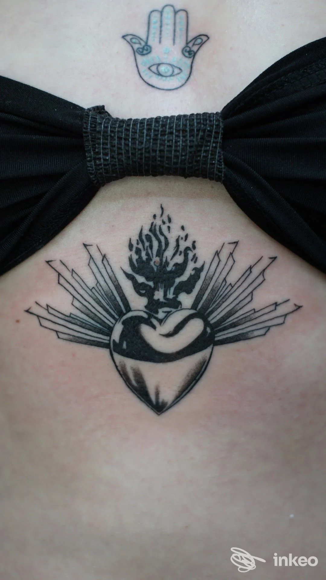 Due tatuaggi sul petto: piccolo hamsa sopra il reggiseno e cuore sacro fiammeggiante sullo sterno. Starskcream Tattoo, Ancona.
