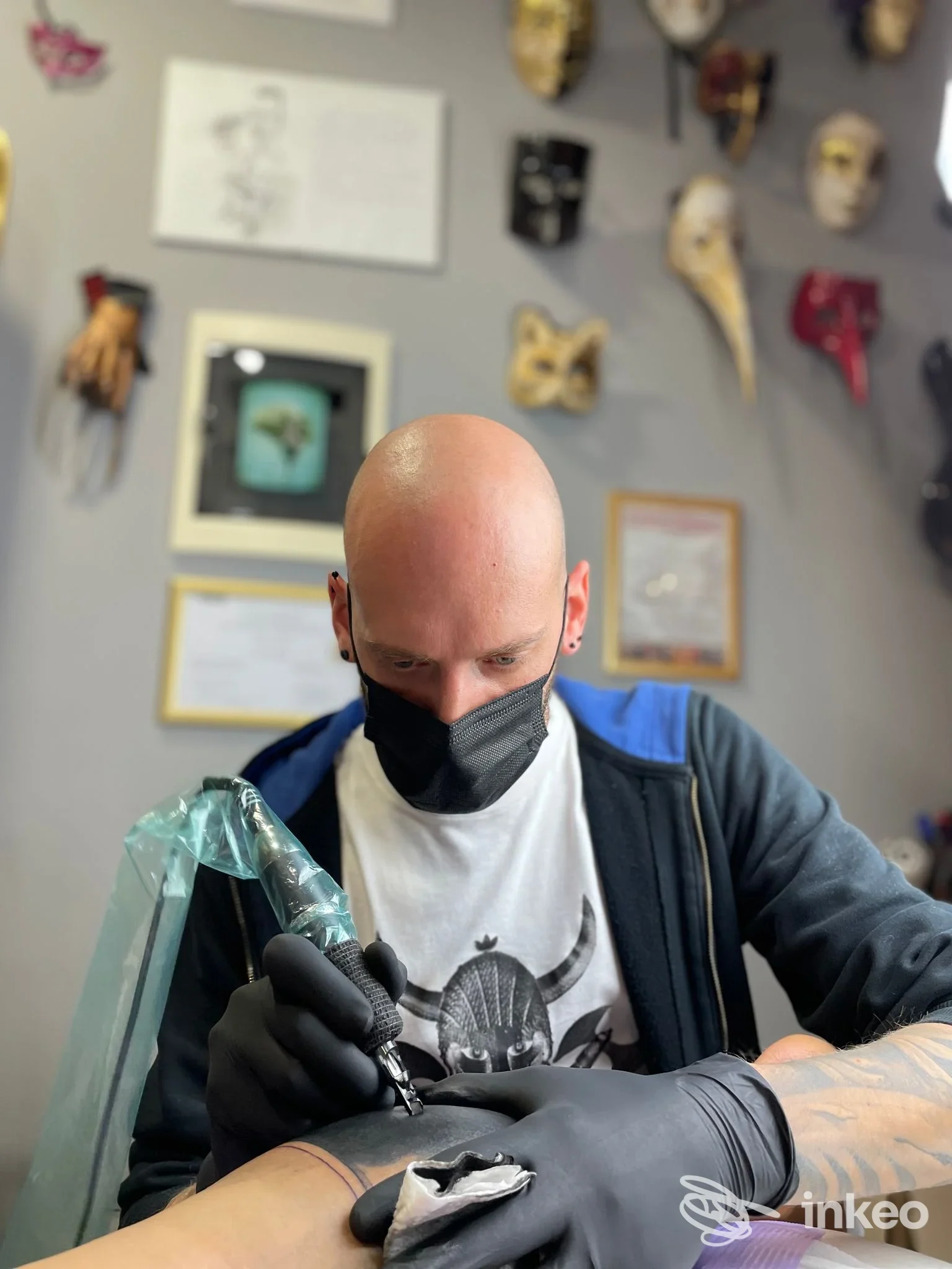 Operatore di Venom Art Tattoo a Rapagnano mentre esegue un tatuaggio sul braccio di un cliente, con maschere decorative sullo sfondo.