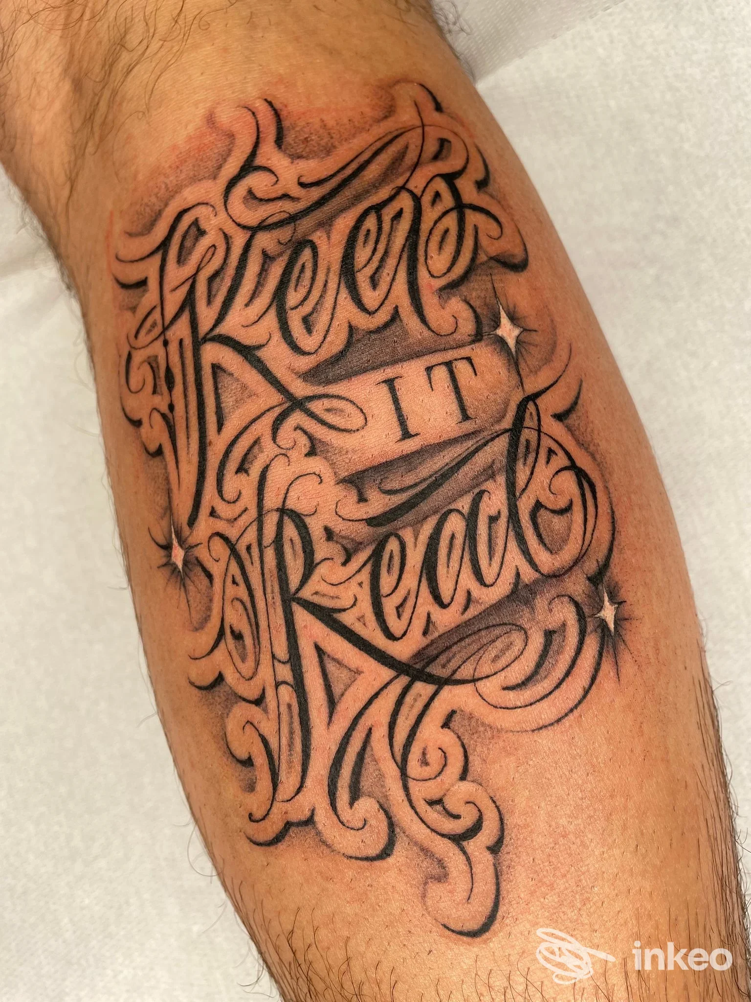 Tatuaggio sul braccio con testo decorativo Keep It Real in stile calligrafico, contorni neri e ornamenti ornamentali.