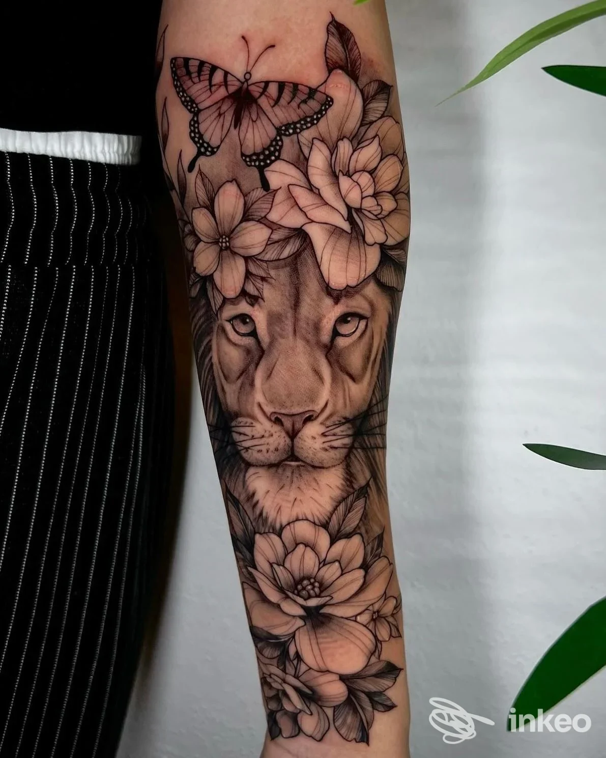 Tattoo realistica sull’avambraccio raffigurante una testa di leone con fiori e una farfalla, in tonalità di nero e grigi.