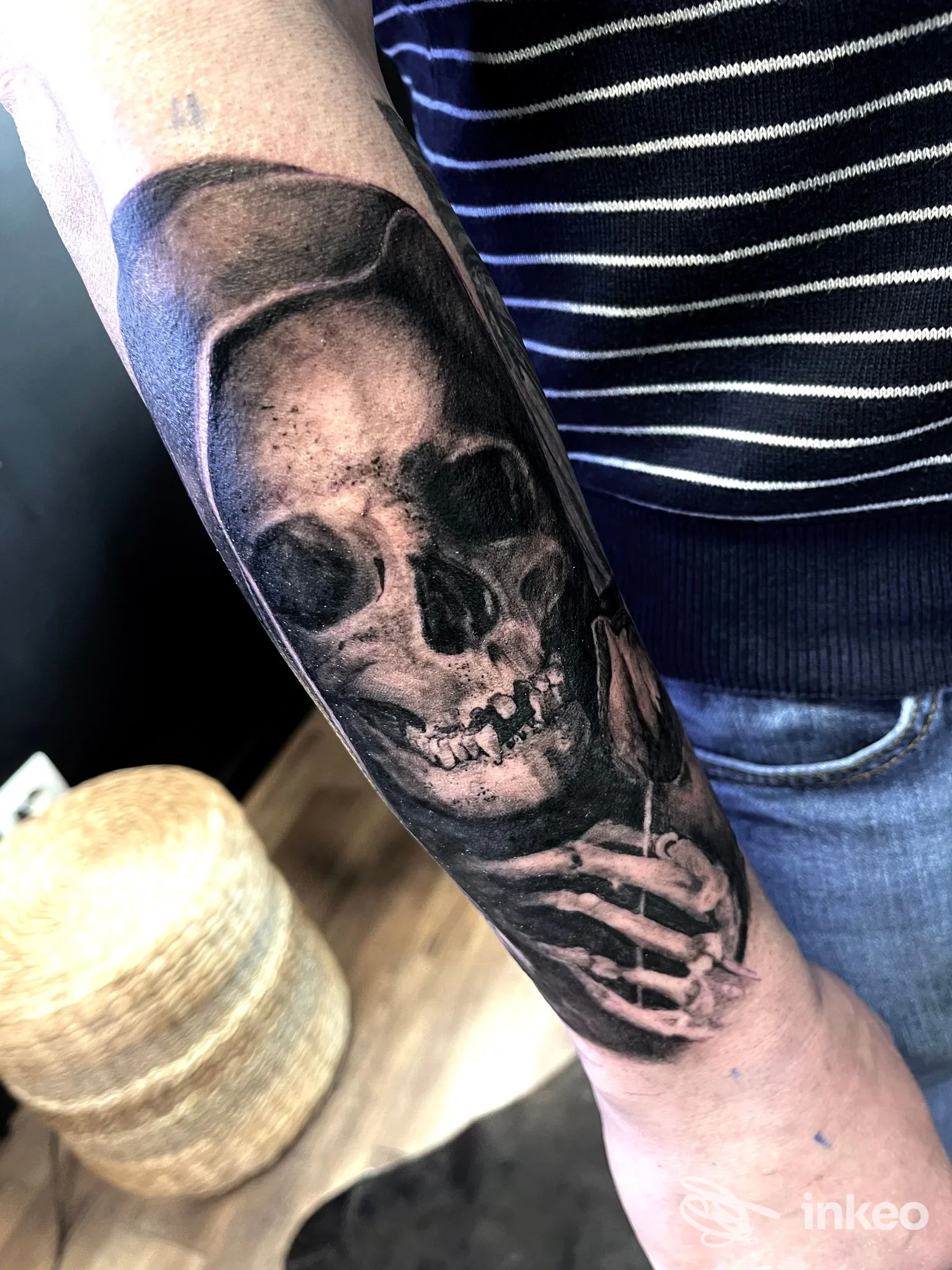 Tatuaggio in bianco e nero di skull e una mano scheletrica sull’avambraccio, visibile una porzione di manica a righe.
