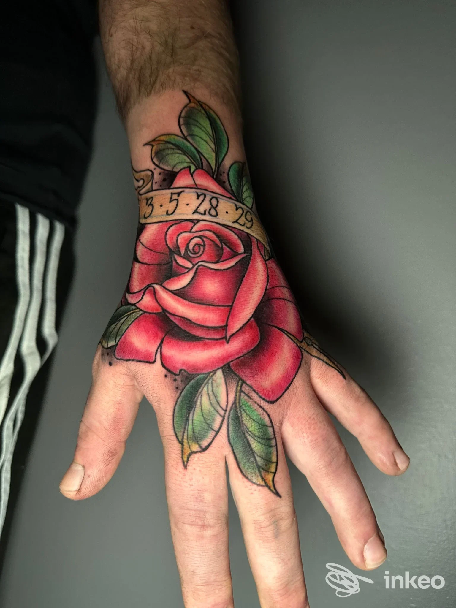 Tatuaggio di una rosa rossa con nastro e numeri sul dorso della mano, scatto dello studio Cecchitatua a Torino, dettaglio a colori.