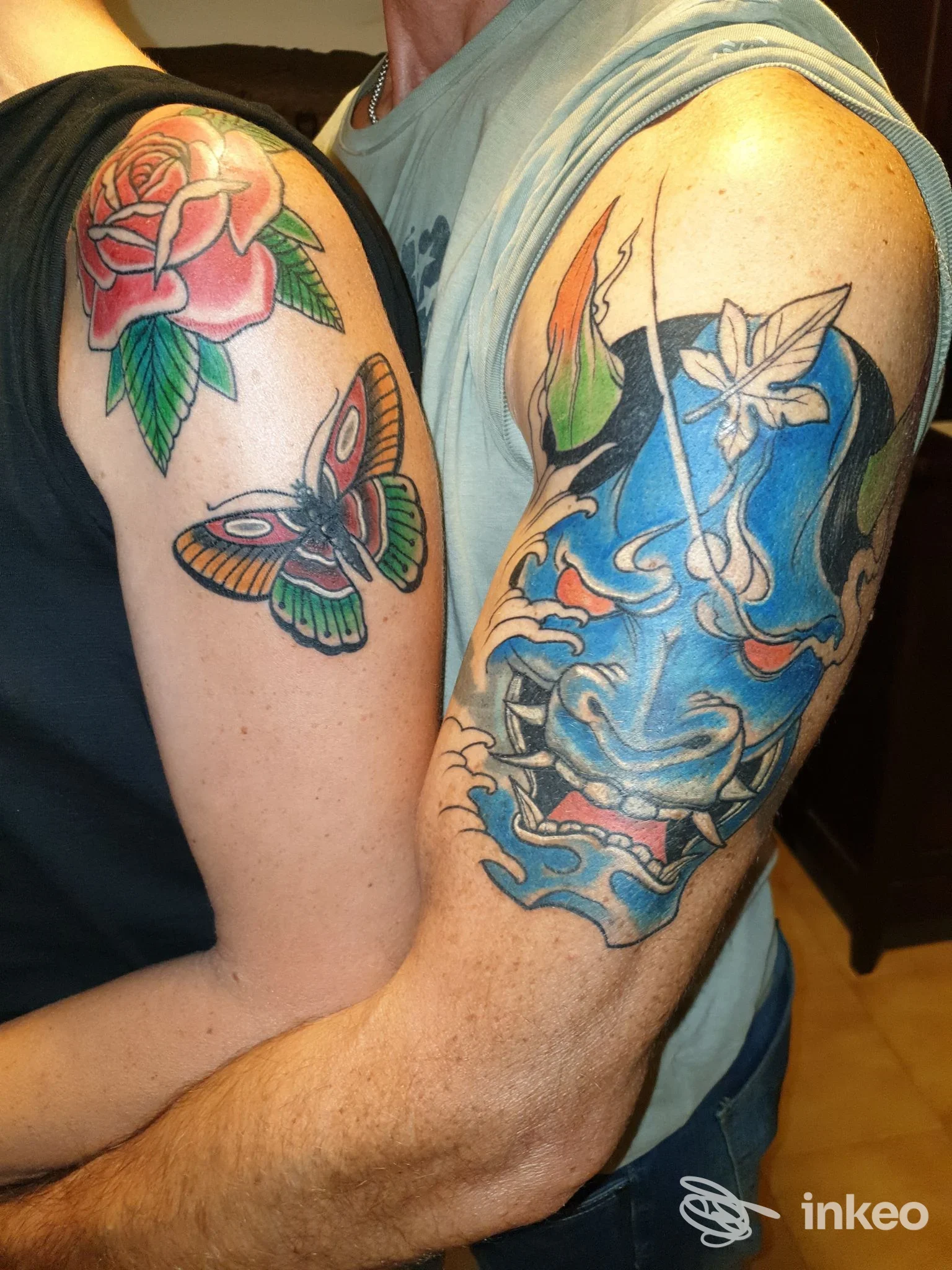 Due braccia con tatuaggi a colori: rosa e farfalla su una spalla, grande maschera blu con fiori su bicipite. Luca Tattoo Story, Volpiano.