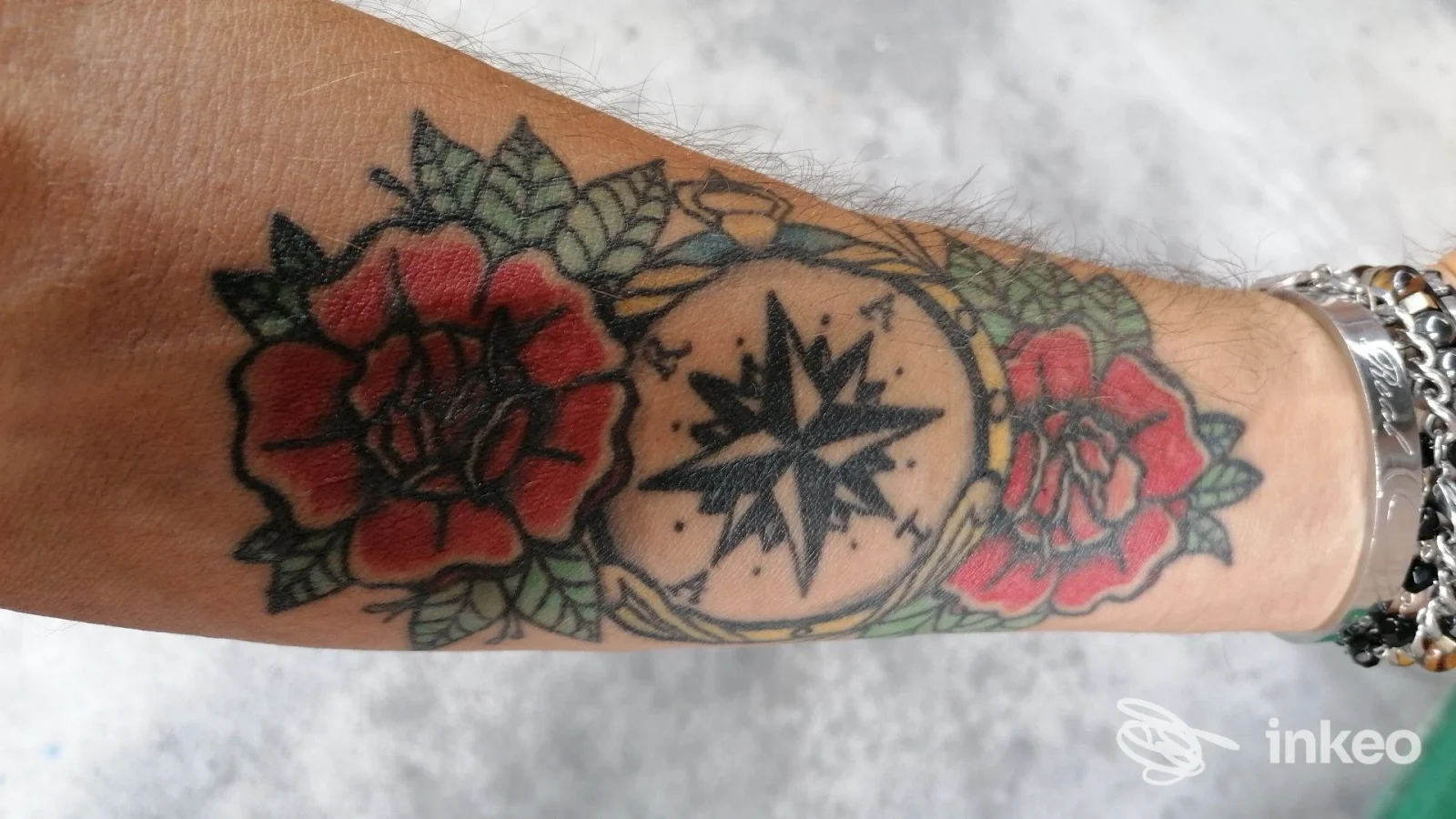 Tatuaggio reale sull'avambraccio: bussola nera centrale circondata da rose rosse e foglie verdi, foto nitida. Malamanera Tattoo Shop, Nichelino.
