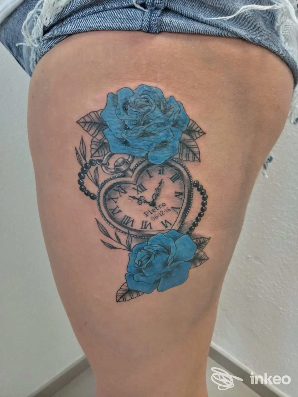 Tatuaggio sul lato coscia con orologio a forma di cuore, catena di perle e rose blu, realizzato da Maxx Tattoo traditional body art a Mondovì.