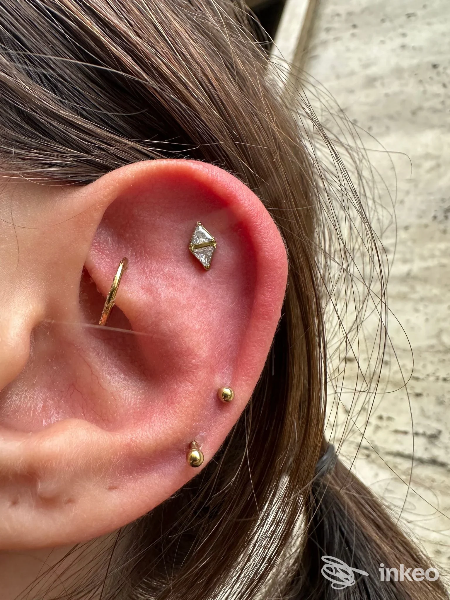 Primo piano di un orecchio con più piercing in oro (helix, tragus e lobo) e un gioiello triangolare, foto per Nuni Piercing Lab Torino, Torino.