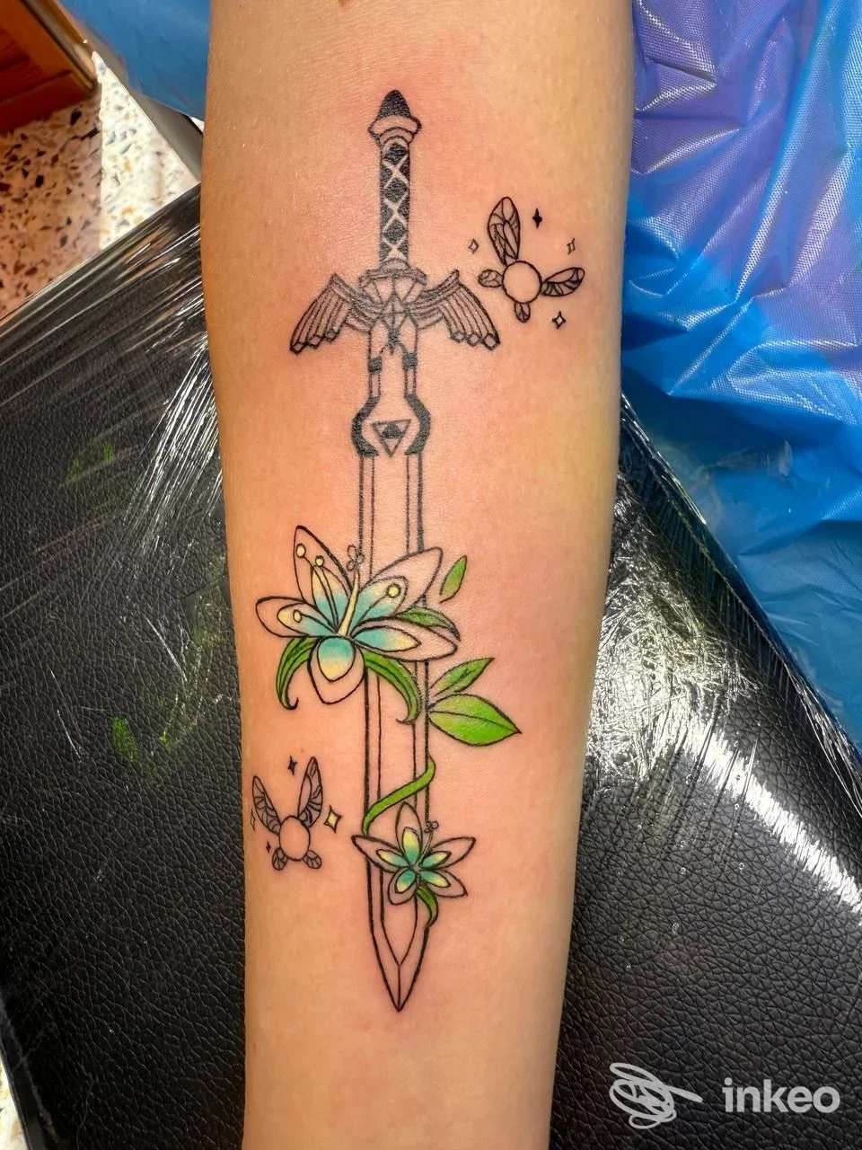 Tatuaggio sull'avambraccio: spada verticale con fiori verdi/azzurri e piccoli elementi volanti, lavoro fotografato da Ritual Tattoo di Ilaria Fachechi, Torino.