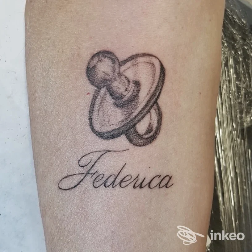 Tattoo su avambraccio raffigurante un ciuccio e la scritta Federica in corsivo.