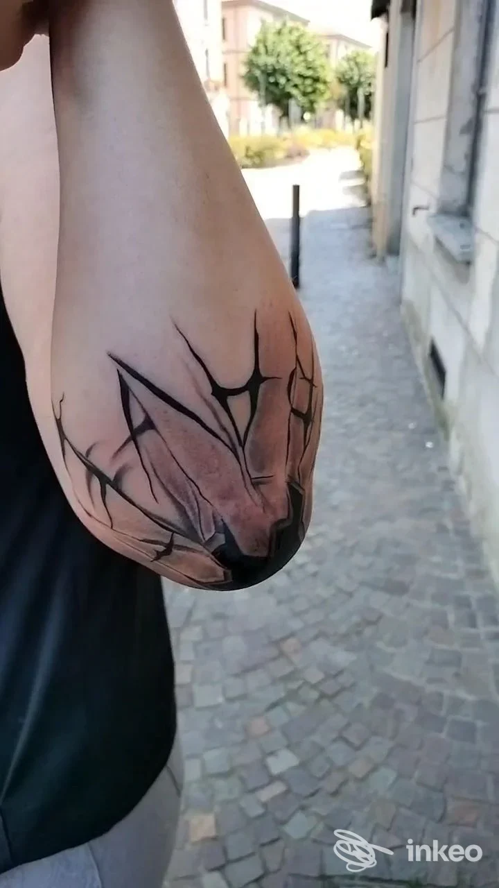 Immagine di un tattoo sul gomito di una persona, linee geometriche nere su pelle chiara, sfondo strada cittadina all'aperto.
