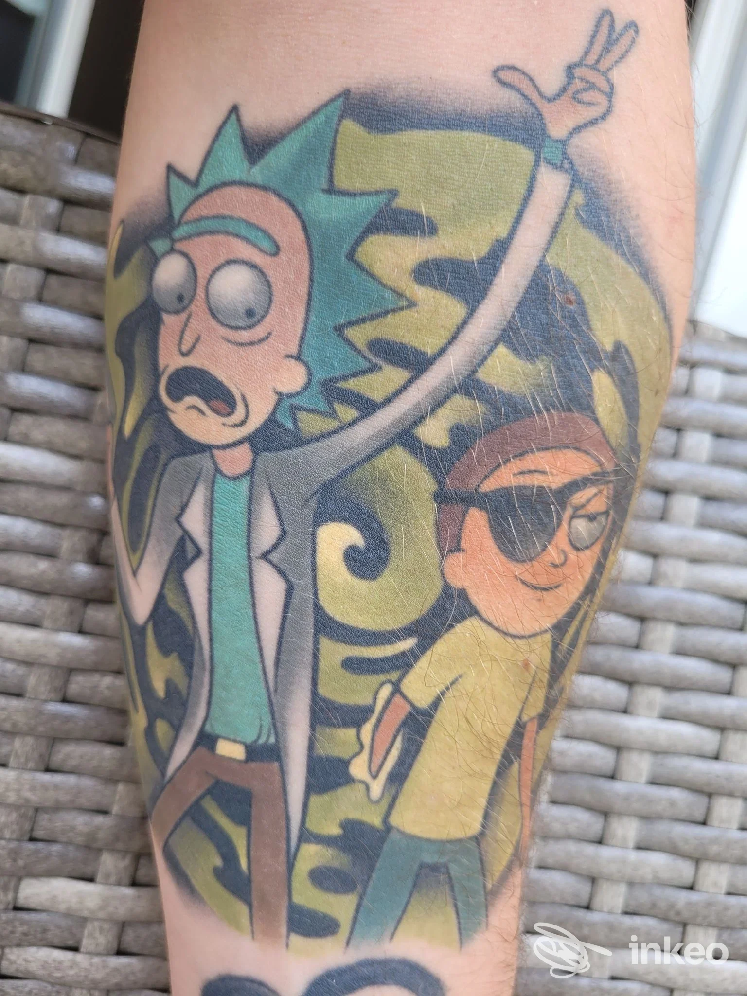 Tatuaggio colorato raffigurante Rick e Morty sull'avambraccio, con sfondo giallo-verde; fotografia per portfolio di The Kingdom Tattoo Studio ad Alessandria.