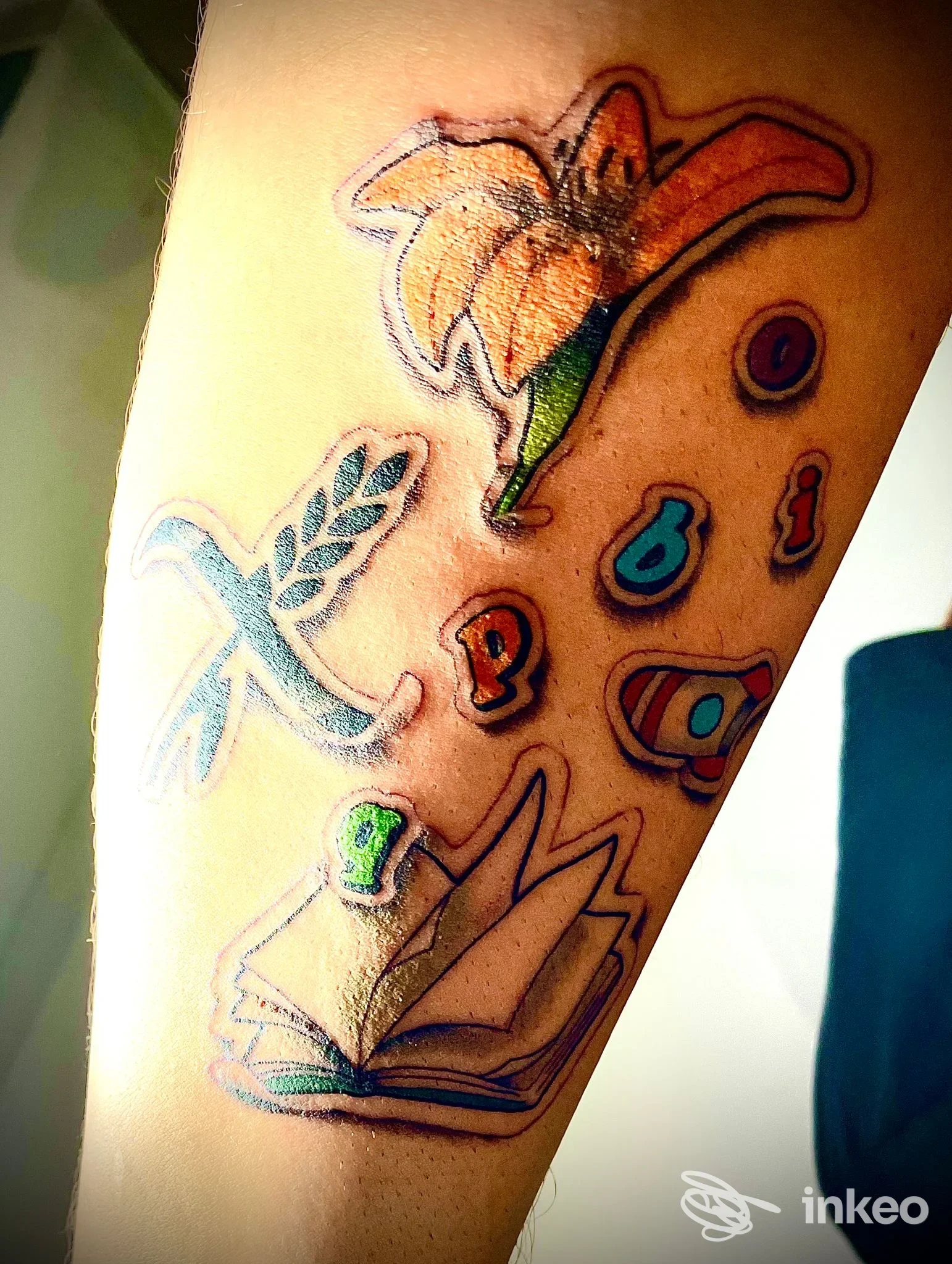 Primo piano di avambraccio tatuato con fiori, libro stilizzato e lettere colorate, immagine fornita da Wild Art Tattoo Studio, Torino.