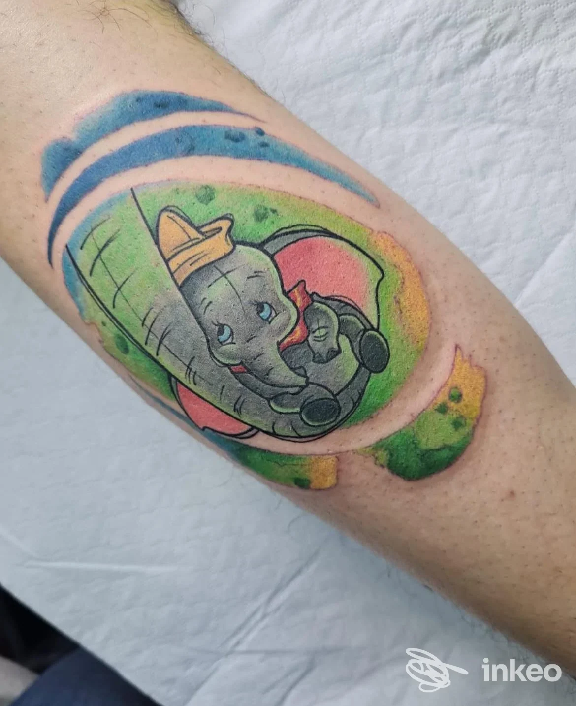 Tattoo colorato di un elefante sul braccio di una persona, contorni definiti e sfondo verde/giallo, senza volto visibile.