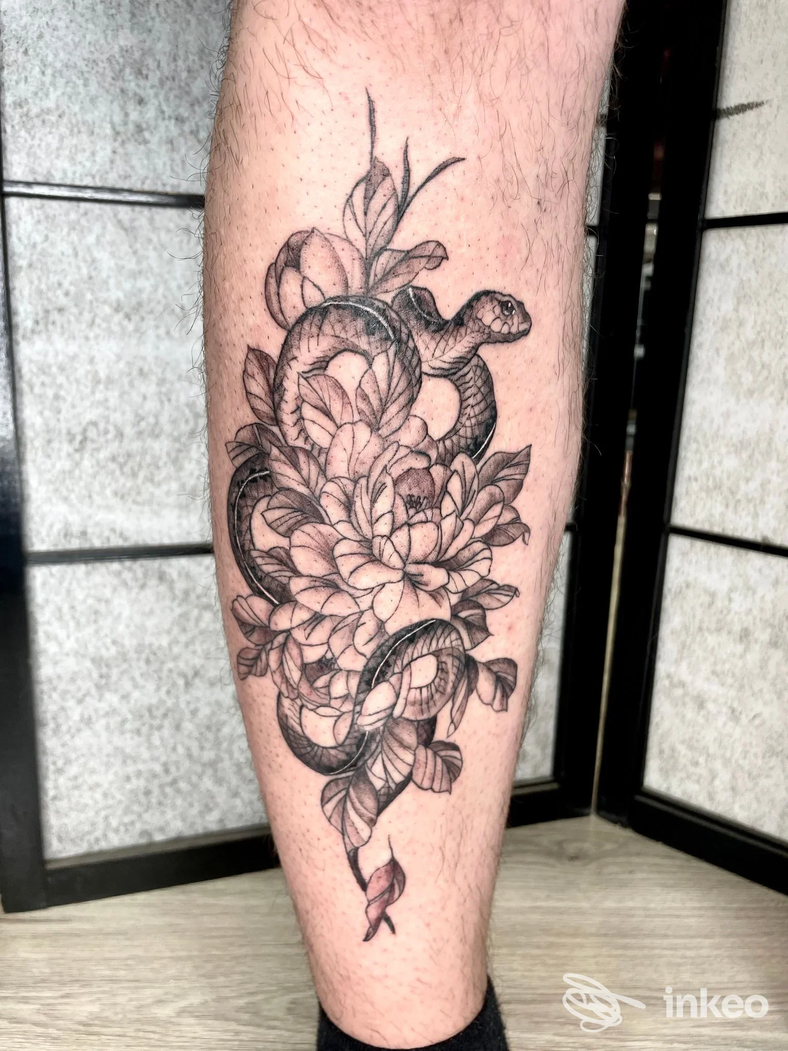 Tatuaggio su polpaccio: serpente avvolto attorno a grandi fiori in bianco e nero, foto fornita da Balam Tattoo Collegno, Collegno.