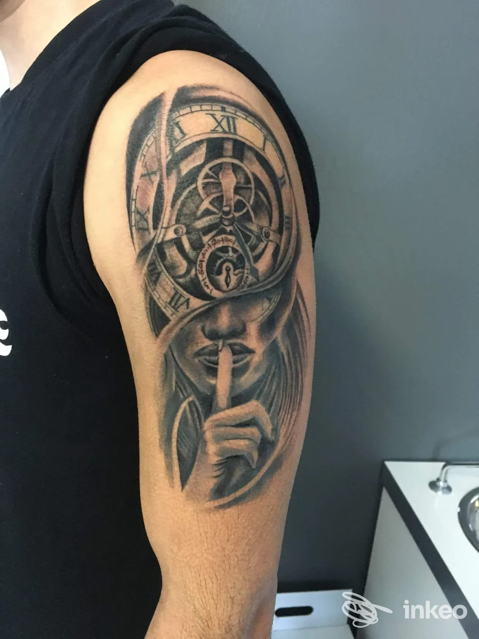 Tatuaggio realistico in bianco e nero su braccio superiore con orologio e volto silenzioso – Fedeli alla Linea Tattoo Studio, Arona