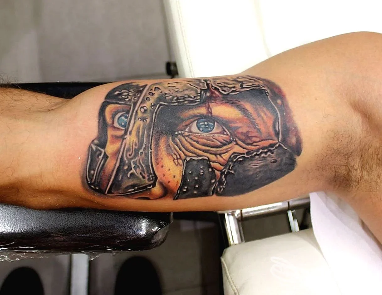 Tatuaggio realistico di volto con elmo sull'interno del bicipite, realizzato da Lele Tattoo Art by Skin Colours ad Alessandria.