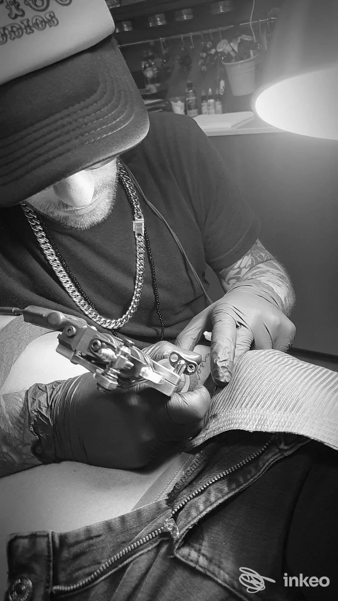 Un tatuatore indossa guanti mentre lavora su una pelle, con una catena al collo; si vede la macchina da tatuaggio in azione sulla pelle del cliente.