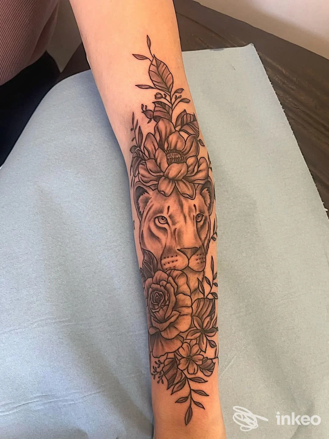Tatuaggio realistico su avambraccio raffigurante testa di leone circondata da fiori e foglie, eseguito da Tattoo Studio by Maury a Chivasso.