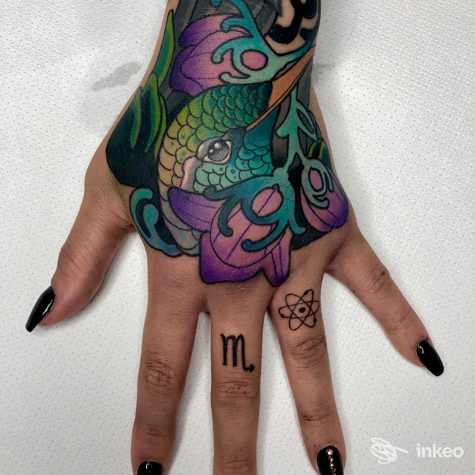Mano con tatuaggio colorato visibile sulla pelle, dito indice decorato con lettering o simbolo piccolo; non appare volto, sfondo neutro a superficie bianca.