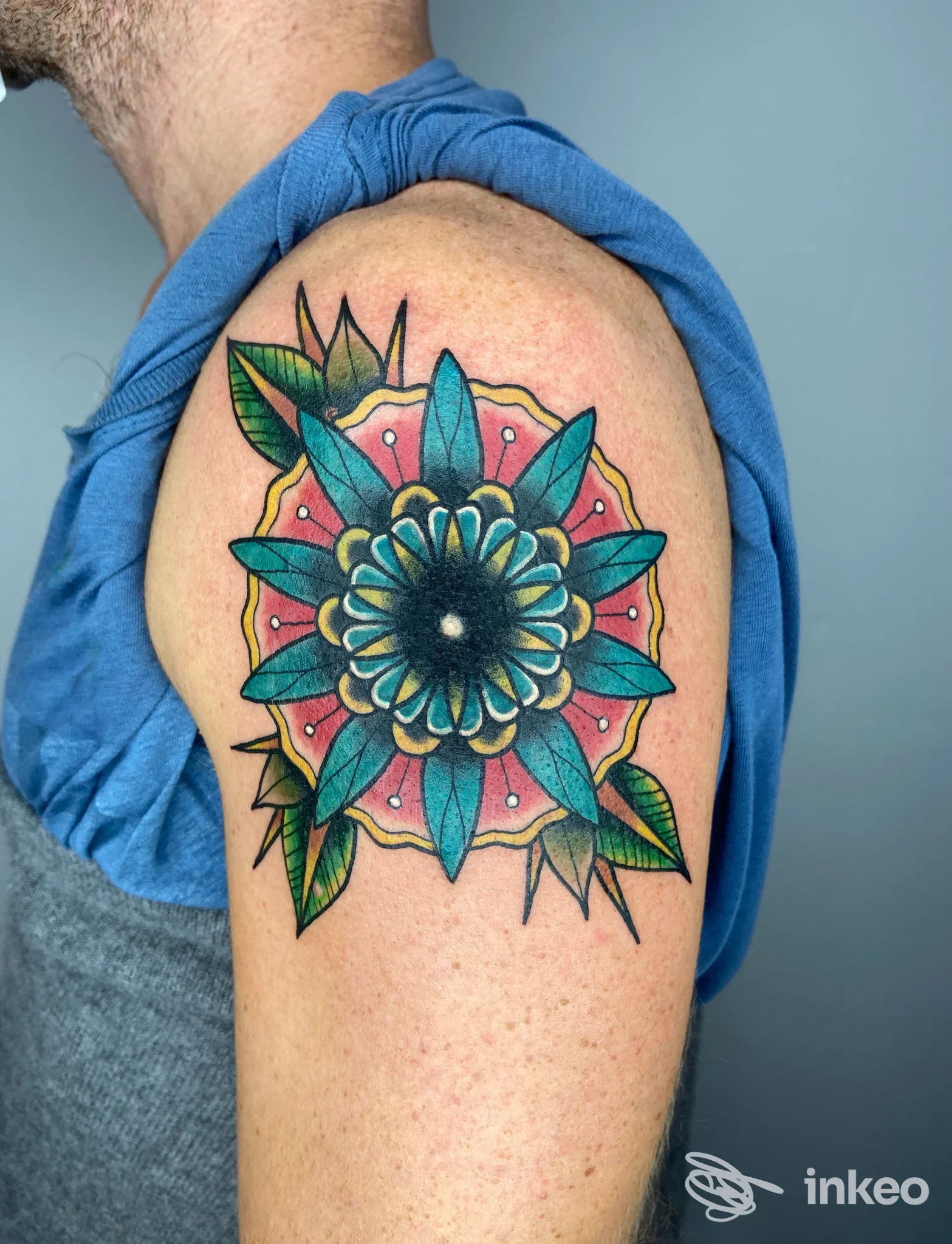Tatuaggio a mandala colorato sulla spalla di una persona, con petali azzurri, verdi e rosa, contorni neri e sfumature, visibile su braccio sinistro. Studio Scighera Tattoo.
