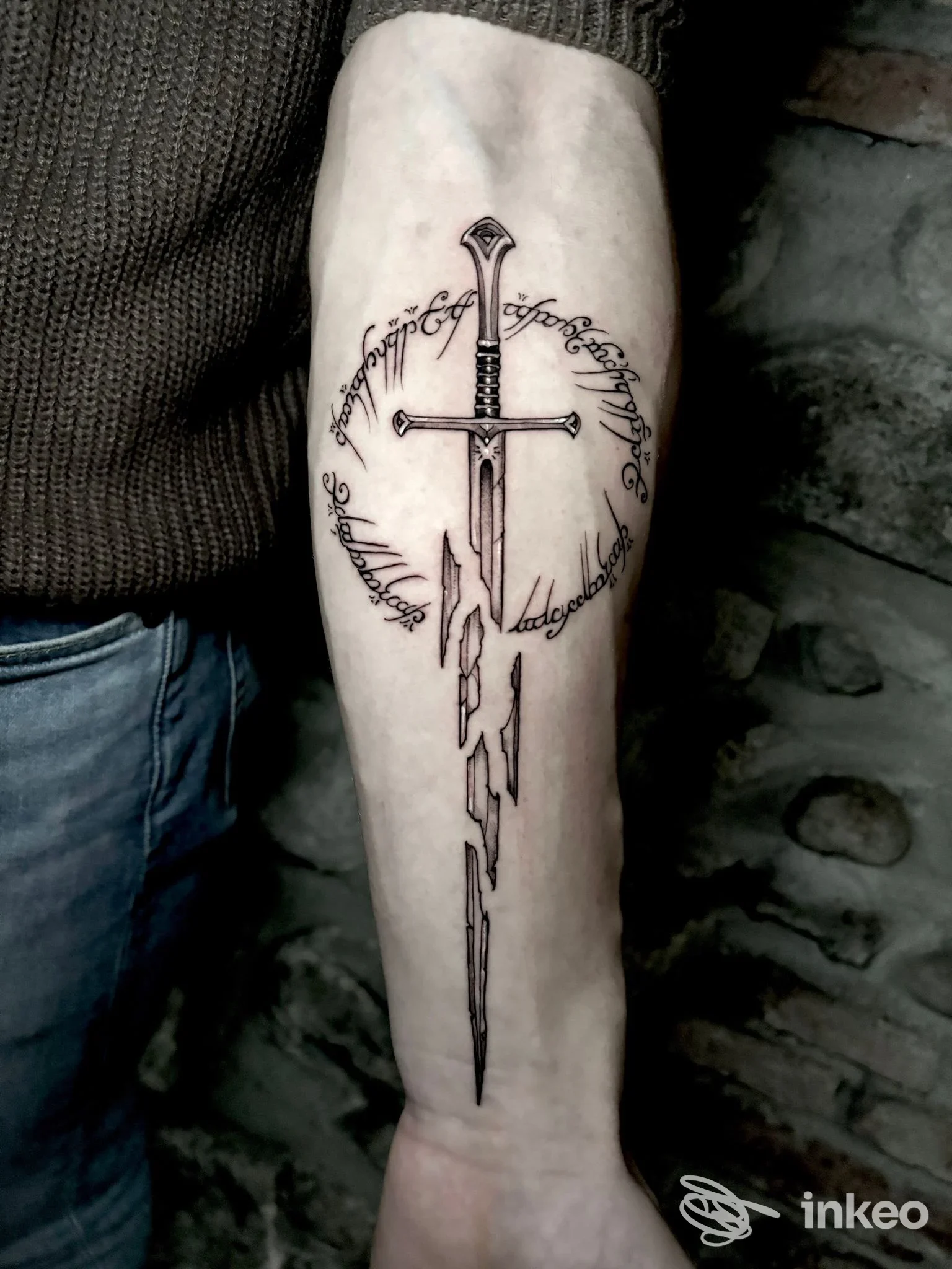 Tatuaggio sull'avambraccio raffigurante una croce con spada centrale, circondato da testo circolare in corsivo, senza segni di overlay né interfacce sociali.