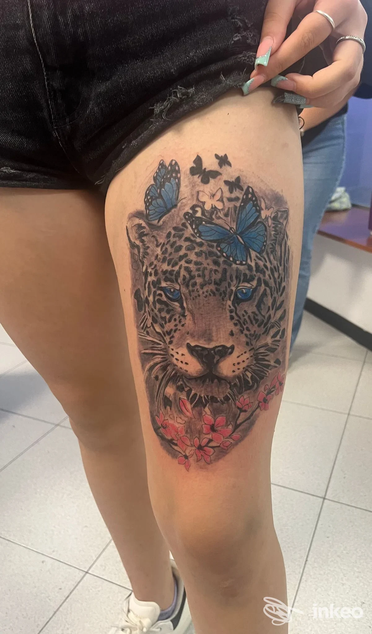 Tatuaggio realistico di leopardo con farfalle blu e fiori rosa sulla coscia, eseguito da Radici Tribali tattoo piercing, Andria.