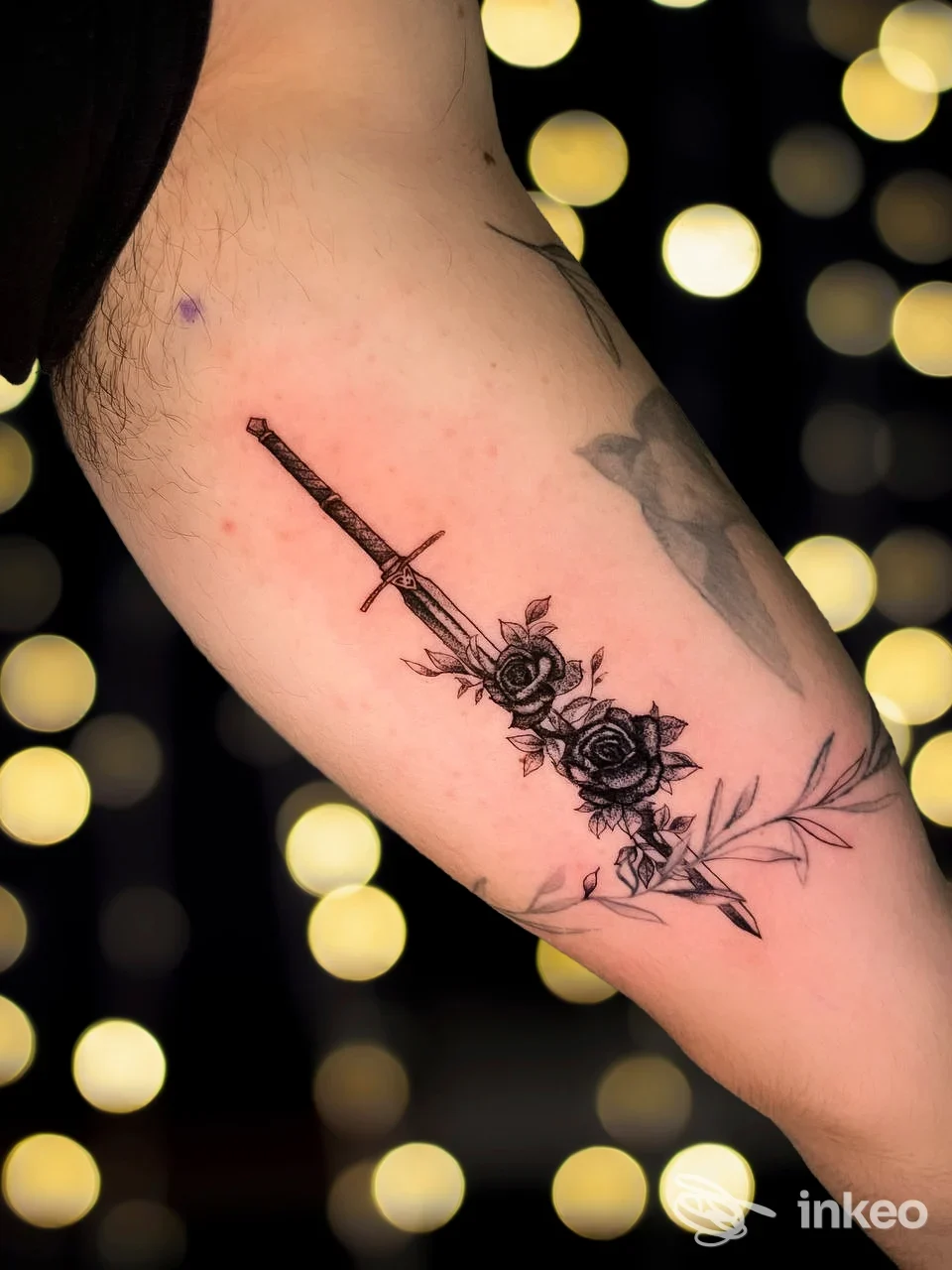 Dettaglio di tatuaggio pugnale con rose sull'interno del braccio, fotografia di Sevenink Tattoo a Taranto con luci bokeh sullo sfondo.