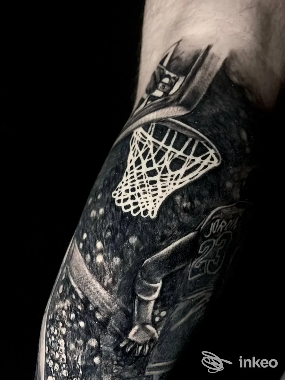 Tattoo in bianco e nero sull'avambraccio raffigurante un canestro da basket e una figura in azione, sfondo scuro e dettagli nitidi.