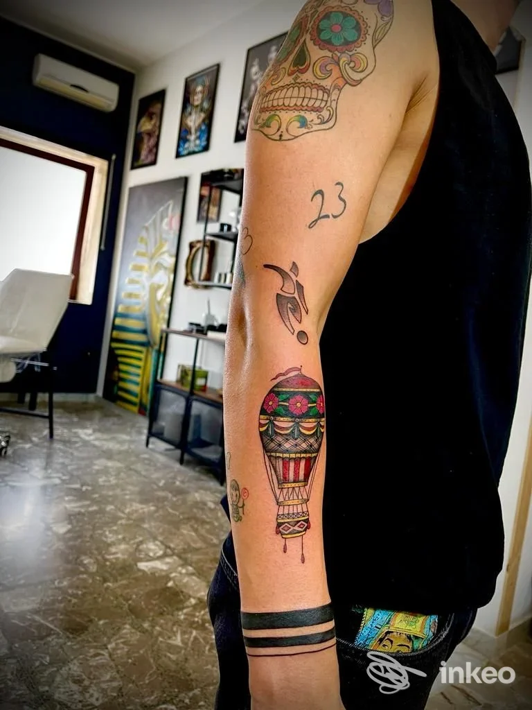 Primo piano del braccio sinistro con più tatuaggi colorati (mongolfiera, fasce nere e teschio) nello studio Tattoo Tankhamon a Torchiarolo.
