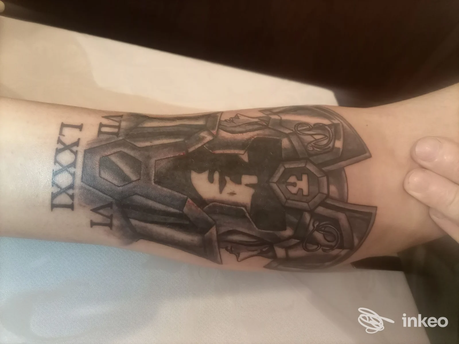 Inchiostro nero su pelle: avambraccio con tatuaggio in stile black and grey che mostra figure geometriche e un volto stilizzato integrato nel disegno; nessun overlay o testo esterno visibile.
