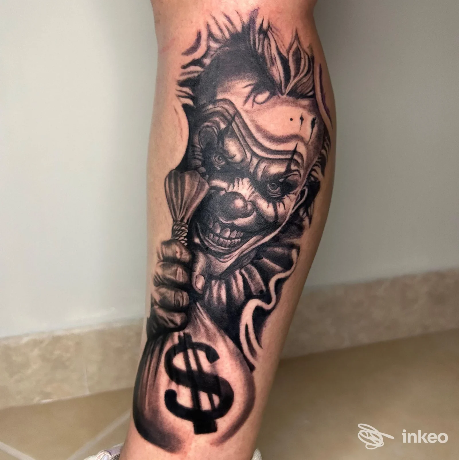 Tatuaggio realistico in bianco e nero di un clown che spunta e tiene un sacco con simbolo del dollaro sul polpaccio. WILD INK TATTOO STUDIO, Martina Franca.