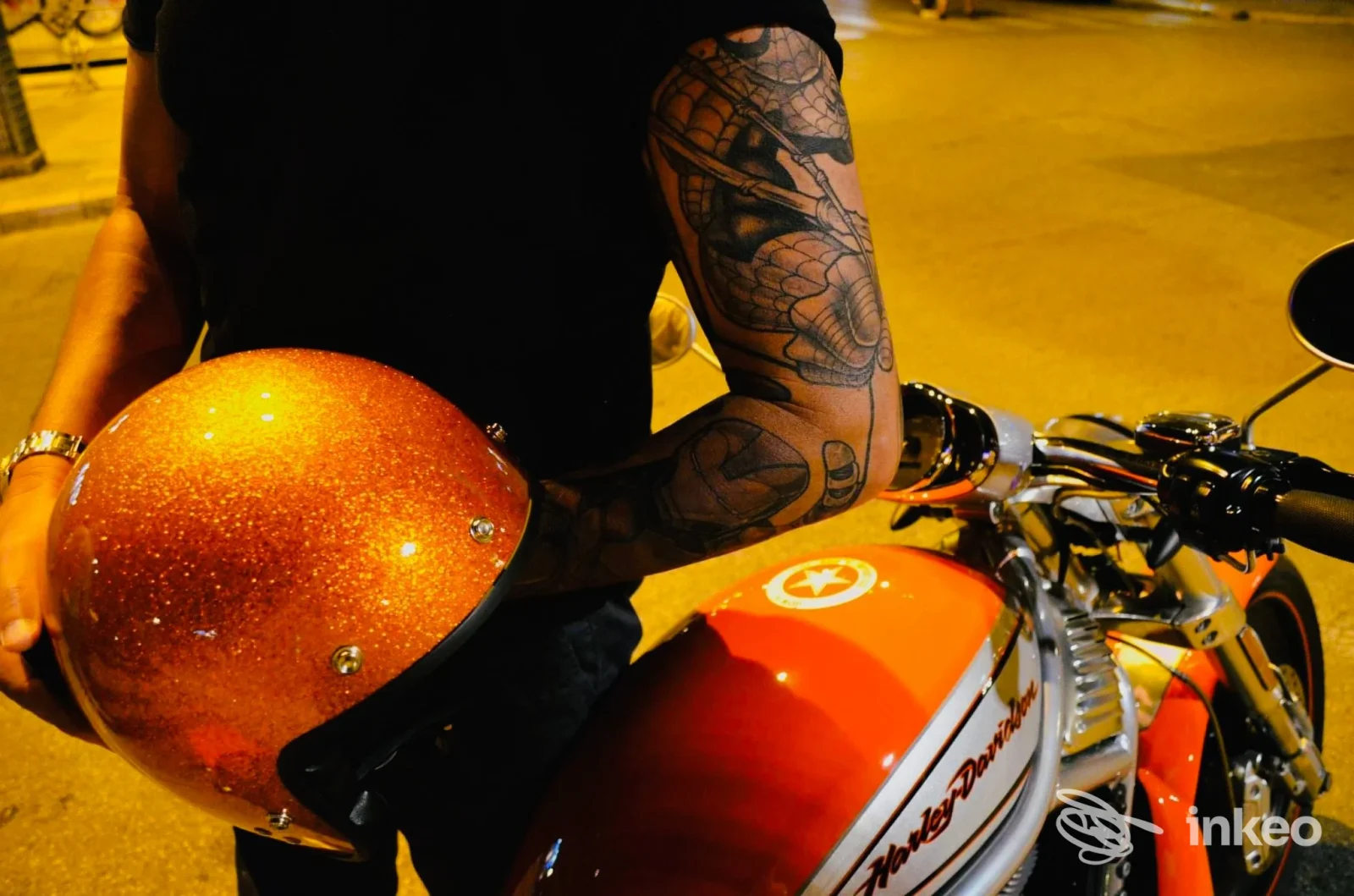 Dettaglio di un avambraccio tatuato mentre la persona tiene un casco glitter arancione, foto scattata accanto a una motocicletta a Bari per Wild Cat Tattoo Studio.
