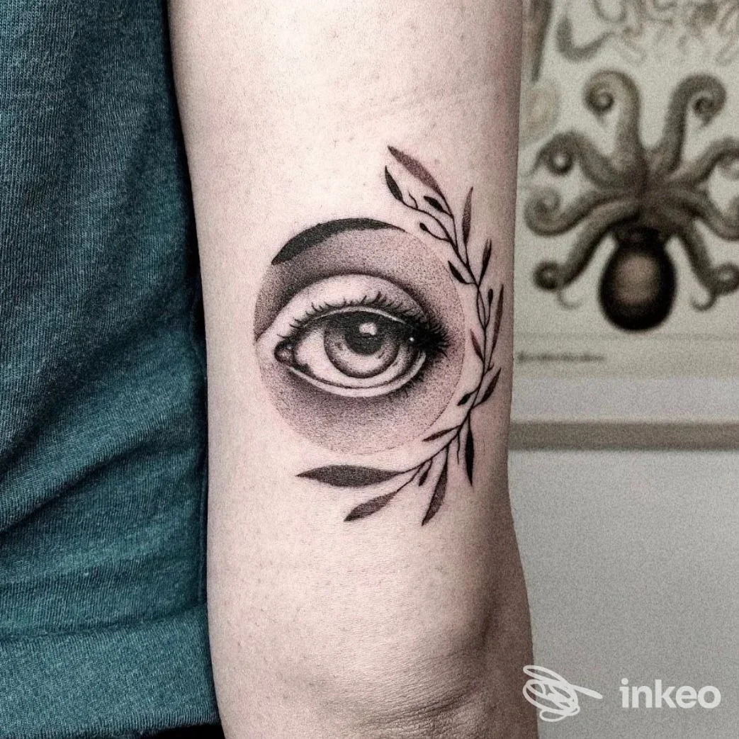 Tattoo realistica di un occhio al centro del braccio, incorniciato da una ghirlanda di foglie scure.
