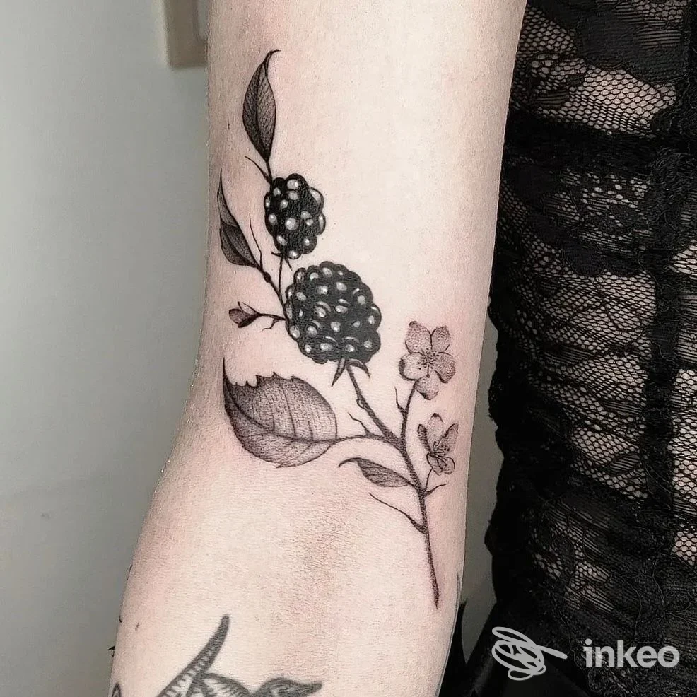 Tattoo botanico su avambraccio: ramo con foglie e bacche scure, fiorellini, in tonalità nere su pelle reale.