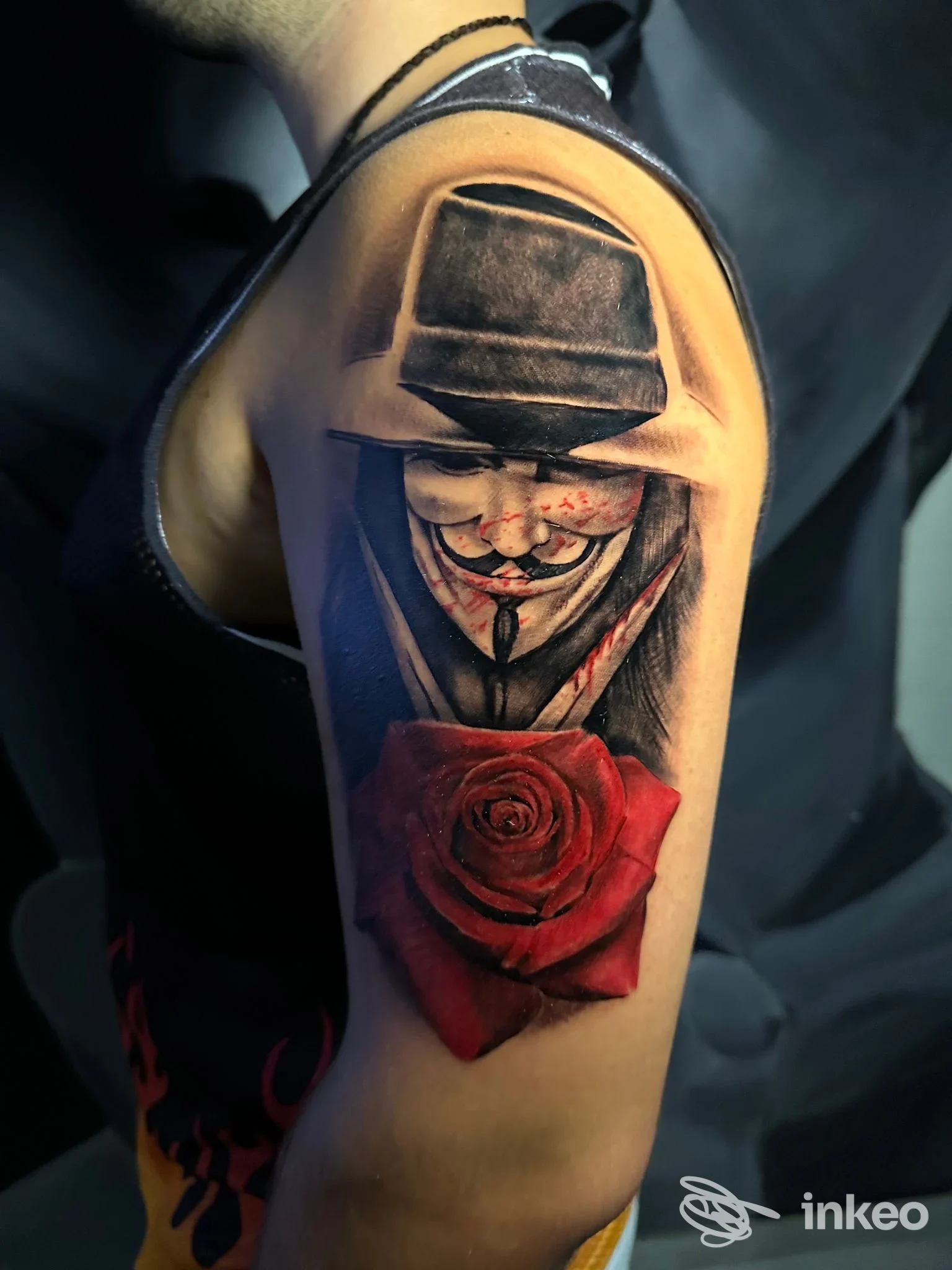 Tatuaggio realistico su braccio: maschera stile Guy Fawkes con segni di sangue e grande rosa rossa, immagine da Arosa Tattoo Studio, Tricase.