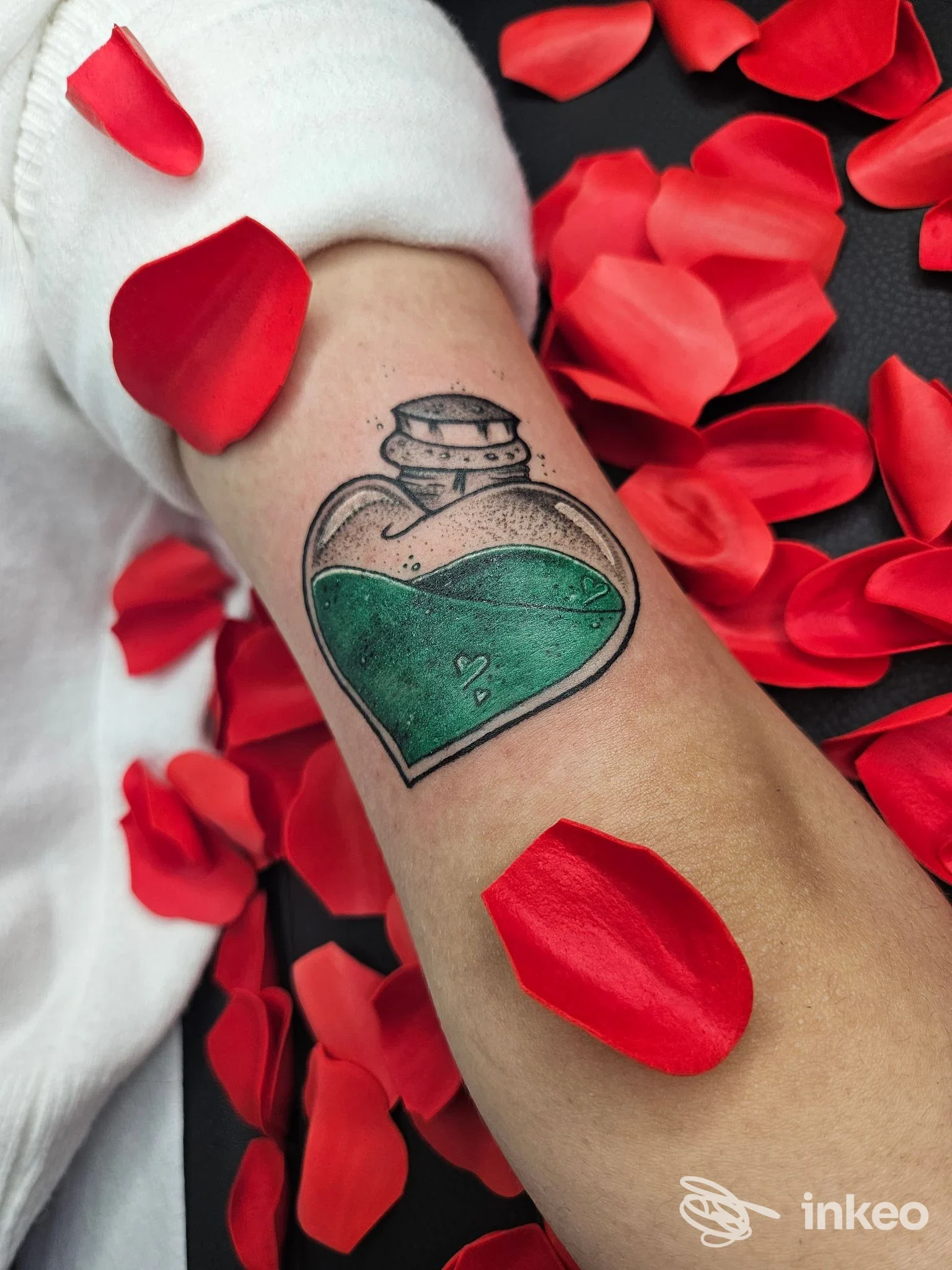 Tattoo sull'avambraccio: cuore a forma di bottiglia di profumo con liquido verde, contorni neri; sfondo di petali rossi, realizzato da Mariposa Tattoo.